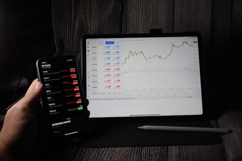 A hand holds a smartphone displaying stock prices next to a tablet showing a financial chart and data.